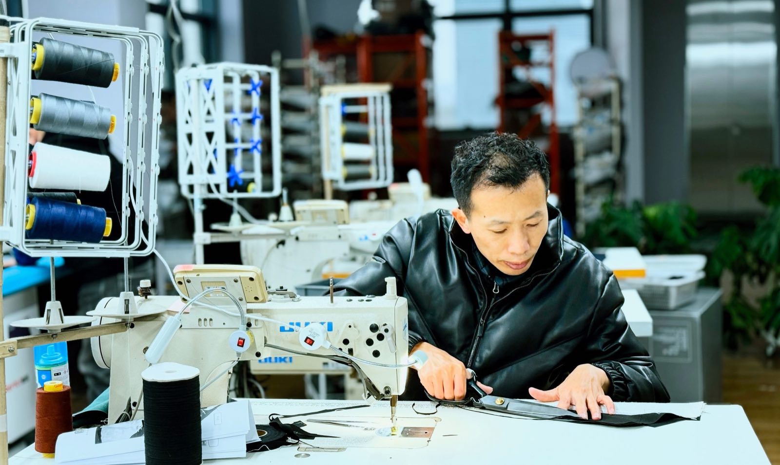 A tailor in the Shanghai workshop marks a cut piece of dark cloth on a table beside thread spools and a sewing machine
